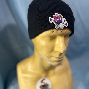May include: Black knit beanie with a colorful paw print patch. The patch features shades of purple, red, and blue. The beanie is displayed on a mannequin head.