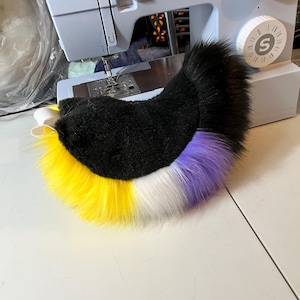 Puede incluir: Un accesorio de cola de piel sintética curvada y afelpada con un diseño de bloques de color que incluye una sección superior negra, seguida de una base amarilla vibrante, una franja central blanca y una sección final morada, con una trabilla elástica blanca.