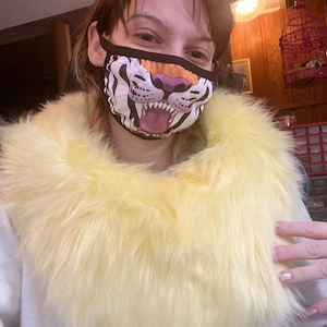 May include: A face mask with a tiger design, featuring a white and black striped pattern, orange nose, and pink mouth. A fluffy yellow collar is around the neck.