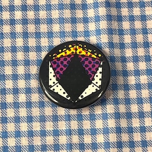 May include: A round, black button with a colorful geometric design. The design features a black diamond shape with purple polka dots, white triangles with white dots, and yellow and orange accents. The button is set against a blue and white checkered background.