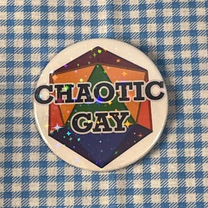 May include: A round button with the text "CHAOTIC GAY" in black letters. The button features a colorful geometric design with orange, green, purple, and blue sections. The button has a holographic, glittery border.