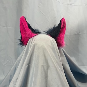 May include: A pair of faux fur animal ears with a headband. The ears are hot pink on the inside and black on the outside, with long, dark fur around the edges. The ears are attached to a silver headband.