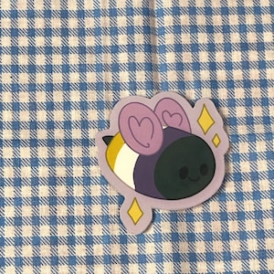 May include: A sticker featuring a cartoon bee with a non-binary pride flag design. The bee has a yellow, white, purple, and black body with heart-shaped wings and a smiling face. The sticker is outlined in purple with yellow diamond accents.