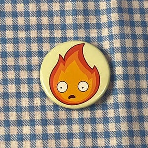 May include: A round button with a cartoon flame design. The flame is orange and red with a surprised expression and white eyes. The button is on a light green background.
