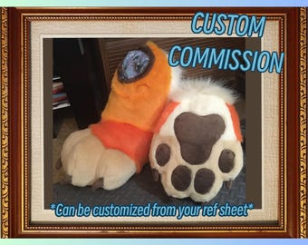 May include: A pair of custom-made furry paws in orange, tan, and white. The paws have a detailed paw pad design with brown pads and white fur accents. The text "CUSTOM COMMISSION" and "Can be customized from your ref sheet" are visible.