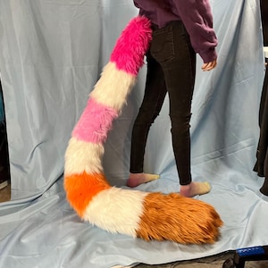 May include: A colorful, faux fur tail in shades of pink, white, orange, and brown. The tail is long and fluffy, with a curved shape. The tail is attached to the back of a person wearing black pants and a purple sweatshirt.
