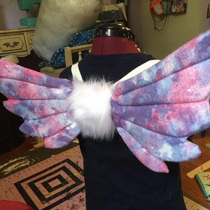 May include: A pair of fantasy-themed wings with a galaxy print in shades of purple, pink, and blue. The wings are attached to a white fur center and have white straps. The wings are displayed on a mannequin.