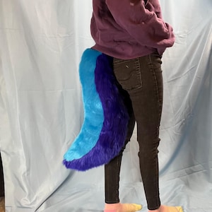 May include: A plush, furry tail in shades of blue and purple. The tail is attached to the back of a person wearing dark jeans and a purple sweatshirt. The tail is a fun accessory.