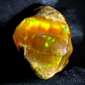 Può includere: Un opale naturale grezzo con un'iridescenza gialla, arancione e verde. L'opale è incastonato in una matrice grigia grezza.