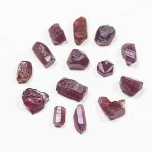 Könnte beinhalten: Eine Sammlung von 13 kleinen, rohen Rubinkristallen in verschiedenen Formen und Größen. Die Kristalle sind tief rotviolett und haben eine natürliche, unpolierte Oberfläche.