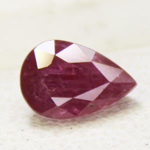 May include: A faceted, pear-shaped ruby gemstone. The gem is a deep, rich red with multiple facets that catch the light. The stone is set against a white background, highlighting its color and shape. This gemstone is suitable for jewelry making.