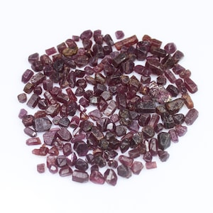May include: A scattering of small, rough-cut ruby gemstones in shades of red and purple. The gemstones are displayed on a white surface, revealing their natural, irregular shapes and textures. The image highlights the raw beauty of the precious stones.