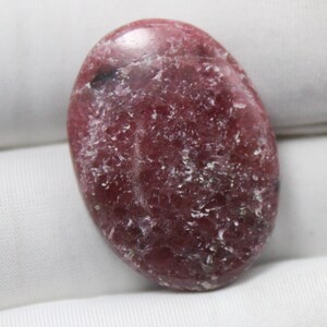 May include: A polished, oval gemstone in shades of deep red and burgundy. The stone has a smooth, glossy surface with subtle white and grey veining. The gemstone is a natural, organic material.