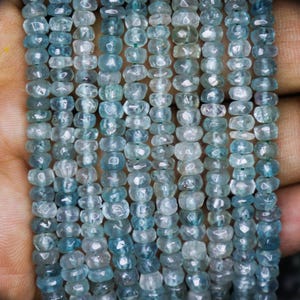 May include: Strands of aquamarine gemstone beads. The beads are a light blue colour and are strung together with a green thread. The beads are small and have a faceted, irregular shape. The beads are displayed against a neutral background.