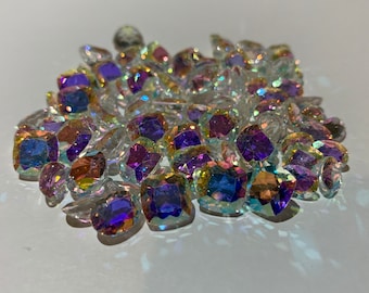May include: A large pile of iridescent, faceted, clear glass gems with purple, blue, and green hues. The gems are scattered on a white surface.