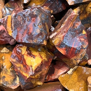May include: A close-up of a pile of red, brown, and yellow jasper stones. The stones are rough and have a natural, unpolished finish.