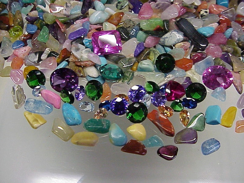 K&ouml;nnte beinhalten: Eine Sammlung von bunten Edelsteinen und Trommelsteinen, darunter Amethyst, T&uuml;rkis und Rubin. Die Steine sind in einem zuf&auml;lligen Muster auf einer wei&szlig;en Oberfl&auml;che angeordnet.