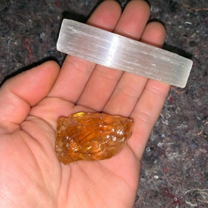 May include: A close-up of a hand holding a small, rough, orange-brown crystal and a long, white, rectangular selenite crystal.