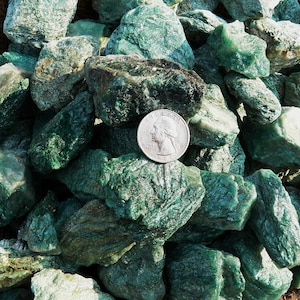 May include: A close-up of a pile of green aventurine rough stones. The stones are various sizes and shapes, with a silvery sheen. A US quarter is included for scale.