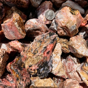 May include: A close-up of a pile of pink, brown, and black stones. The stones are rough and have a variety of textures. A silver coin is visible in the upper left corner of the image.