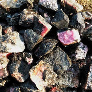 May include: A pile of rough, dark gray and pink rhodonite stones. The stones are irregularly shaped and have a natural, unpolished finish.
