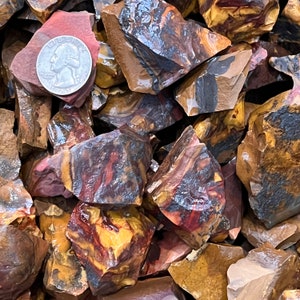 May include: A close-up of a pile of red, brown, and yellow banded agate stones. The stones are rough and have a natural, unpolished finish. A US quarter is included for scale.