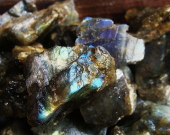 May include: A close-up of a pile of rough, raw labradorite crystals. The crystals are a dark gray color with iridescent blue and green flashes.