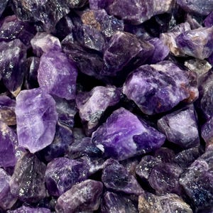 May include: A close-up view of a pile of rough, natural amethyst crystals. The crystals are a deep purple color and have a variety of shapes and sizes.