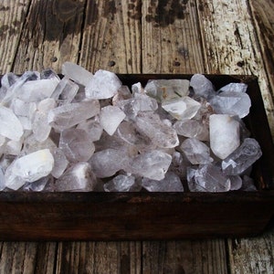 May include: A wooden box filled with clear quartz crystals. The crystals are various sizes and shapes, and some have a slight cloudy appearance.