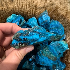May include: A collection of rough, vibrant blue-green mineral stones. The stones have a crystalline appearance with dark, textured inclusions. The stones are displayed in a burlap sack, creating a natural and earthy aesthetic.