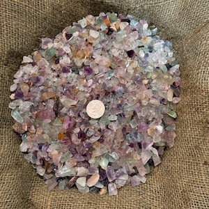 May include: A large pile of colorful fluorite crystals in various shapes and sizes. The crystals are a mix of purple, green, and white, and they are arranged in a burlap sack.