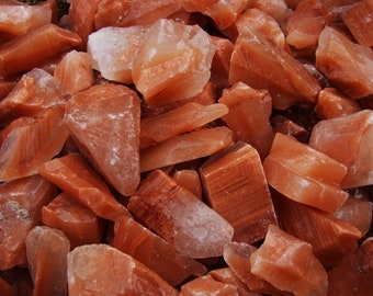 May include: A close-up of a pile of Himalayan pink salt crystals. The crystals are various sizes and shapes, with a reddish-orange hue.