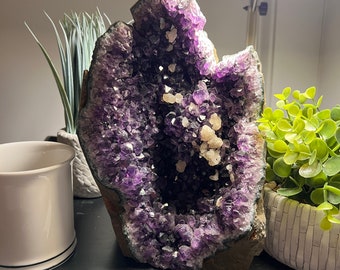 May include: A large amethyst geode with a deep purple color and a natural, rough exterior. The geode is open, revealing a hollow interior lined with sparkling crystals.
