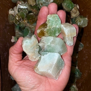 May include: A hand holding a collection of green and white crystal stones. The stones are rough and have a natural, unpolished look.