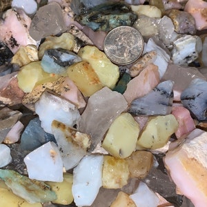 May include: A collection of mixed color, rough-cut gemstones. The stones are various shades of pink, white, green, and yellow. A US quarter is included for scale.