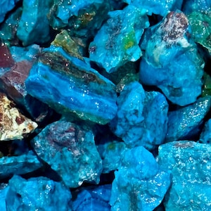 May include: A close-up of a pile of rough, vibrant blue-green chrysocolla stones. The stones have a crystalline structure with areas of brown and tan. The image is well-lit, highlighting the natural textures and colors of the gemstones.