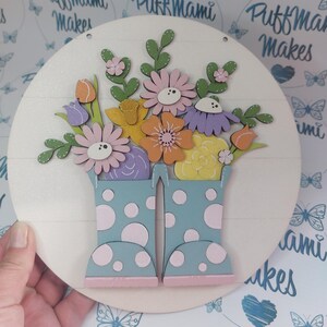 May include: A circular wooden sign with a spring design. The sign has a light background with horizontal lines. A pair of blue wellington boots with white polka dots holds a bouquet of colourful wooden flowers. The flowers include pink, orange, yellow, and purple blooms.