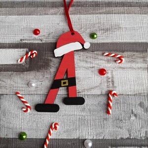 May include: Red wooden Christmas ornament shaped like the letter A, with a Santa hat and a black belt. The ornament is surrounded by candy canes, red and green beads, and white pearls on a woodgrain background.