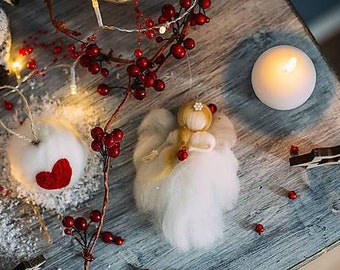 Puede incluir: Adorno de ángel de fieltro blanco con un corazón rojo y un halo dorado, rodeado de bayas rojas y una vela blanca sobre una superficie de madera.