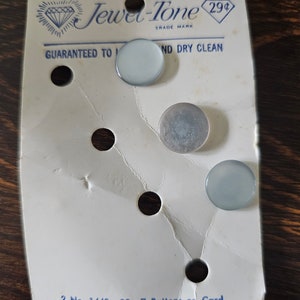May include: A card with four buttons on it. The card is white with blue text that reads "Jewel-Tone 29¢ Trade Mark Guaranteed to... and Dry Clean 2 No. 1440-22-7 Buttons on Card Made in U.S.A. 486". There are three white buttons and one silver button.