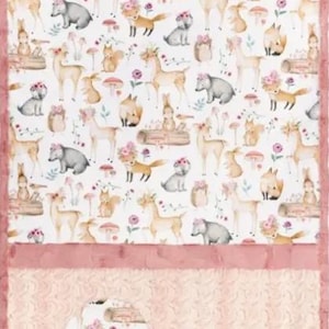 May include: A baby blanket with a whimsical forest animal pattern. The white background features deer, foxes, bears, and other creatures with floral accents. The blanket has a pink border and a textured bottom section.