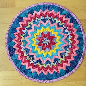 May include: A round, handmade quilted trivet with a vibrant starburst design. The trivet features a central star in yellow, surrounded by pink, blue, and teal zig-zag patterns. A lavender border frames the design, creating a colorful and decorative kitchen accessory.