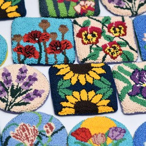 May include: A collection of colorful, handcrafted, floral-themed coasters. Designs include sunflowers, poppies, irises, and lavender, with a variety of shapes and backgrounds. The coasters are made with a textured, looped fabric.