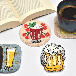 May include: A collection of handmade, embroidered coasters. Designs include a beer bottle and glass, a cocktail, and a beer mug. A mug of coffee sits on a matching coaster. An open book is in the background.