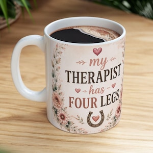 以下が含まれることがあります： 花柄の縁取りと茶色の文字で「my THERAPIST has FOUR LEGS」と書かれた白いセラミック製マグカップ。デザインには小さなピンクのハート、蹄鉄のアイコンが含まれており、マグカップの中には濃い色の液体が入っています。
