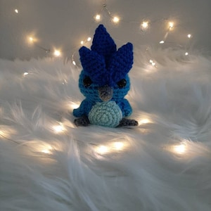May include: A blue and green crocheted bird toy with a spiky blue crest. The toy is sitting on a white fluffy surface with white string lights.