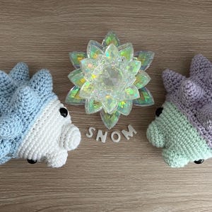 May include: Two crocheted plushies shaped like a spiky Pokemon, one blue and white, the other purple and green, with the word "SNOM" spelled out in white letters between them.