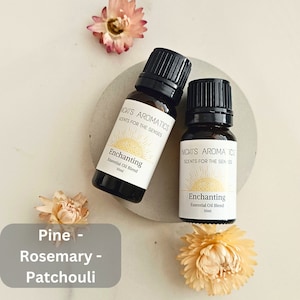 May include: Two 10 ml amber glass bottles of essential oil blend labelled "Enchanting" with a sun graphic and the text "Nicki's Aromatics Scents for the Senses". The bottles are on a white surface with dried flowers.
