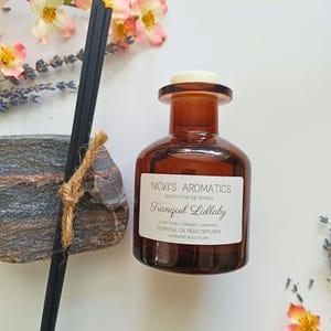 May include: A brown glass bottle with a white label that reads "NICKI'S AROMATICS SCENTS FOR THE SENSES Tranquil Lullaby Clary Sage | Lavender | Geranium ESSENTIAL OIL REED DIFFUSER HANDMADE IN SCOTLAND". The bottle is sitting on a white surface.