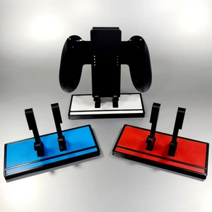 May include: Three Nintendo Switch game console controllers stand on a white, blue, and red base. The controllers are black and the bases are made of plastic.